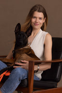 Hey Honey woman in white knit vest and jeans holds a Malinois puppy in a black leather chair.