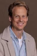 Smiling man in a light beige jacket and blue denim shirt for Hey Honey.
