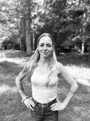Hey Honey, a smiling woman with long blonde hair and tattoos, wearing a tank top and jeans, stands outdoors.