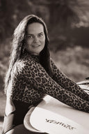 Hey Honey, smiling woman in leopard print rash guard with wet hair, holding a white surfboard.