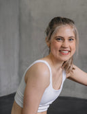 Smiling woman in white Hey Honey sports bra, blonde ponytail, blue eyes, gray background.