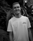 Hey Honey Adrian smiling in a white "yoga at home club" t-shirt, dark tropical foliage background.